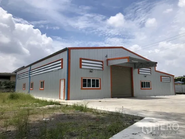 Warehouse for rent in Mueang Nonthaburi, Nonthaburi - Front exterior of a new steel-clad warehouse with orange trim, roller shutter door and wide concrete yard under a bright sky.