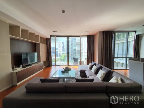 Condominium for rent in Khlong Toei, Bangkok - Spacious living room with L-shaped sofa, built-in TV wall and floor-to-ceiling windows opening to a city-view balcony.