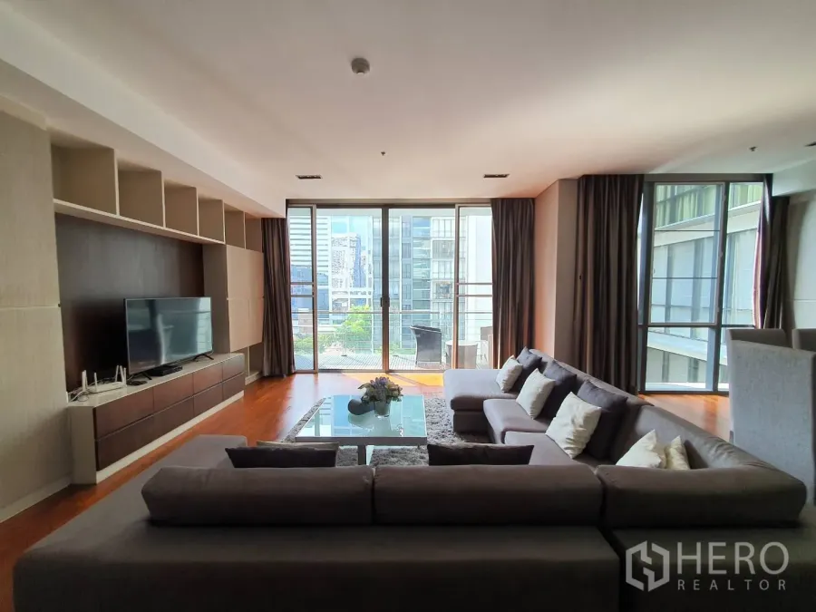 Condominium for rent in Khlong Toei, Bangkok - Spacious living room with L-shaped sofa, built-in TV wall and floor-to-ceiling windows opening to a city-view balcony.