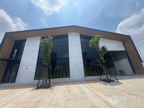 货栈 出租在Bang Sao Thong, Samut Prakan - 现代仓库外立面，超高玻璃窗、景观树木与划线停车位，天空晴朗。