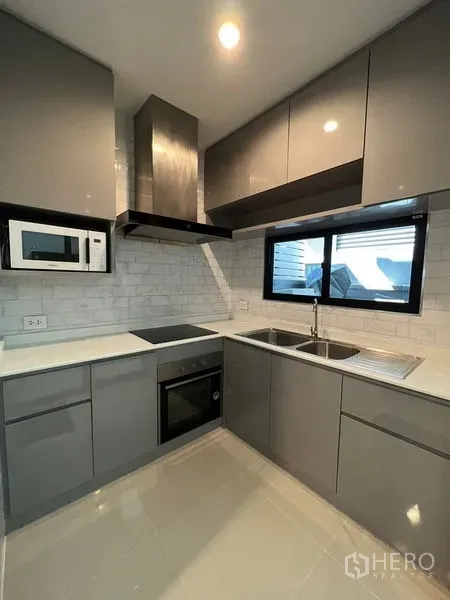 Townhouse for rent in Suan Luang, Bangkok - Sleek L-shaped kitchen featuring grey cabinets, oven, cooktop, hood, sink and subway tile backsplash.