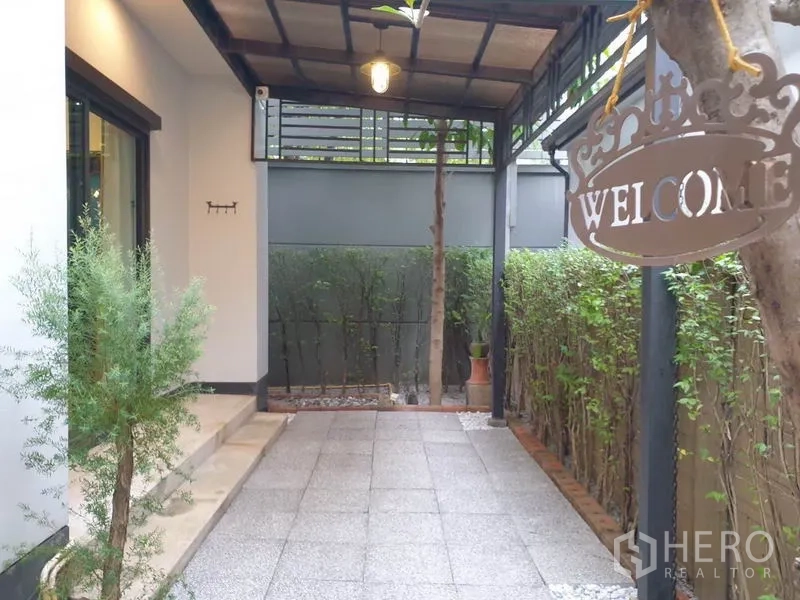 บ้าน ให้เช่าในBang Kapi, Bangkok - ลานนั่งเล่นมีหลังคา พื้นกระเบื้อง ขอบรั้วพุ่มไม้ และป้ายต้อนรับแขวน.