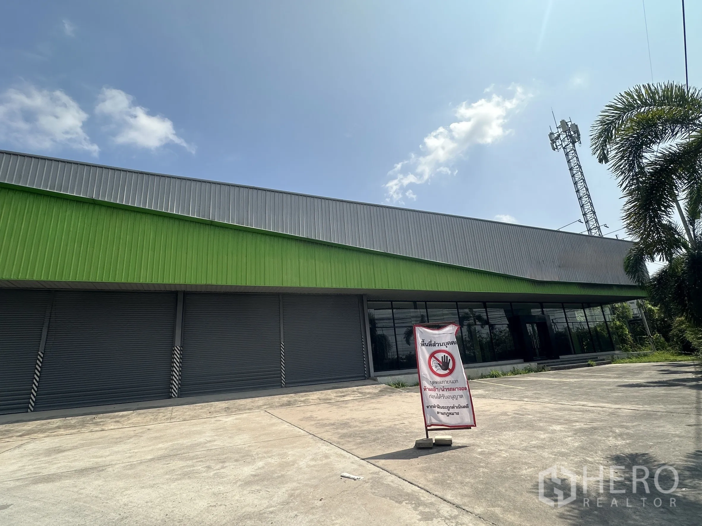 โชว์รูม for rent in Bang Pakong, Chachoengsao - มุมมองเฉียงของโชว์รูมพร้อมผนังกระจก ช่องประตูม้วน ลานจอดกว้าง และเสาโทรคมนาคมกับต้นปาล์มใกล้เคียง
