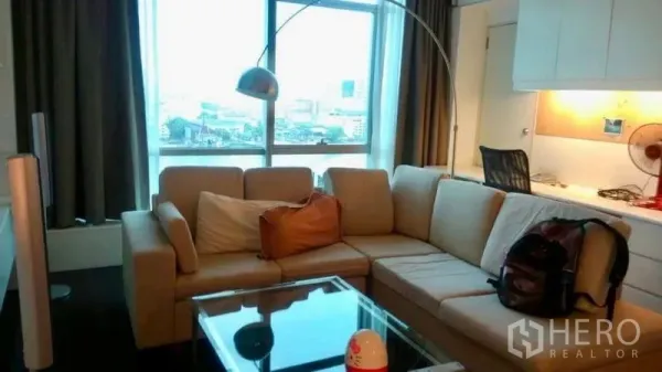 Condominium for sale in Bang Rak, Bangkok - Cozy living room with L-shaped sofa, glass coffee table, arc floor lamp and city view window.