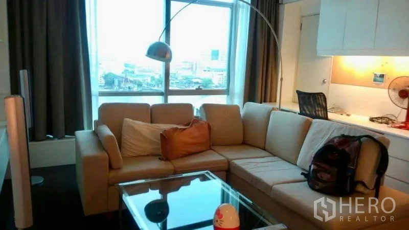 Condominium for sale in Bang Rak, Bangkok - Cozy living room with L-shaped sofa, glass coffee table, arc floor lamp and city view window.