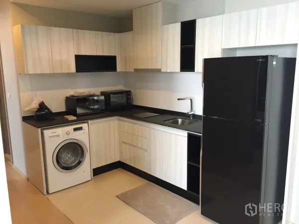 Condominium for rent in Watthana, Bangkok - L-shaped kitchen with light wood cabinets, black countertop, fridge and front-load washing machine.
