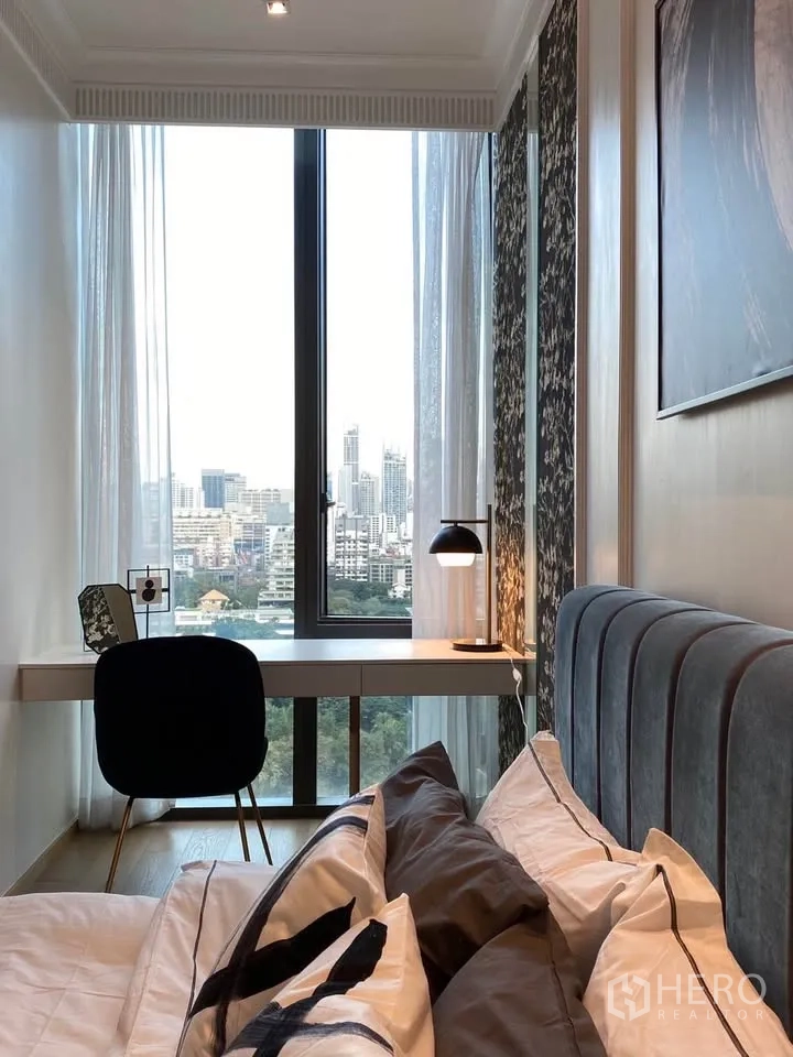 Condominium for rent in Pathum Wan, Bangkok - Bedside view of the built-in desk and lamp facing a floor-to-ceiling window with skyline scenery.
