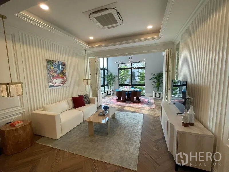 บ้าน ให้เช่าหรือขายในSaphan Sung, Bangkok - ห้องนั่งเล่นครอบครัว โซฟาสีขาว ตู้วางทีวี และประตูบานพับเปิดสู่ซันรูมกระจก