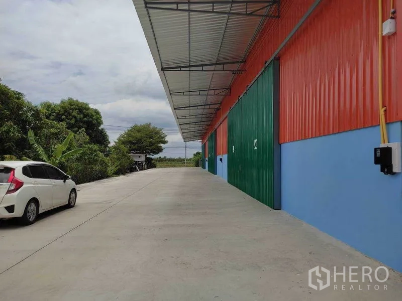 คลังสินค้า ให้เช่าในBang Yai, Nonthaburi - ทางวิ่งคอนกรีตกว้างข้างอาคารโกดังผนังส้ม-ฟ้า ประตูบานเลื่อนสีเขียว พร้อมกันสาด