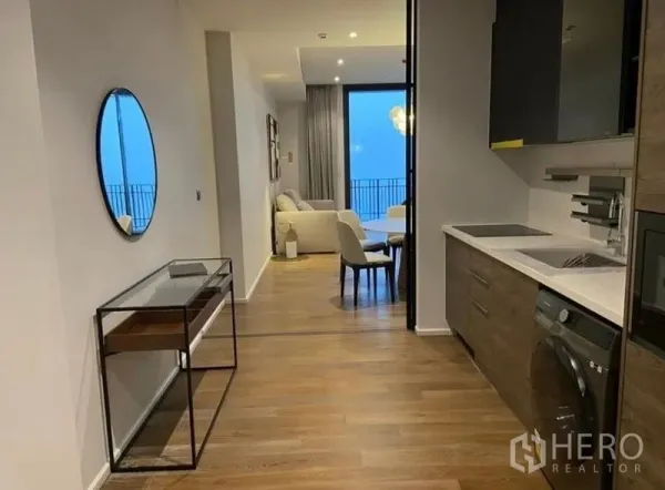 Condominium for rent in Pathum Wan, Bangkok - Hallway view to a bright living area with a fitted galley kitchen featuring induction hob, oven, washer, and wood floors.