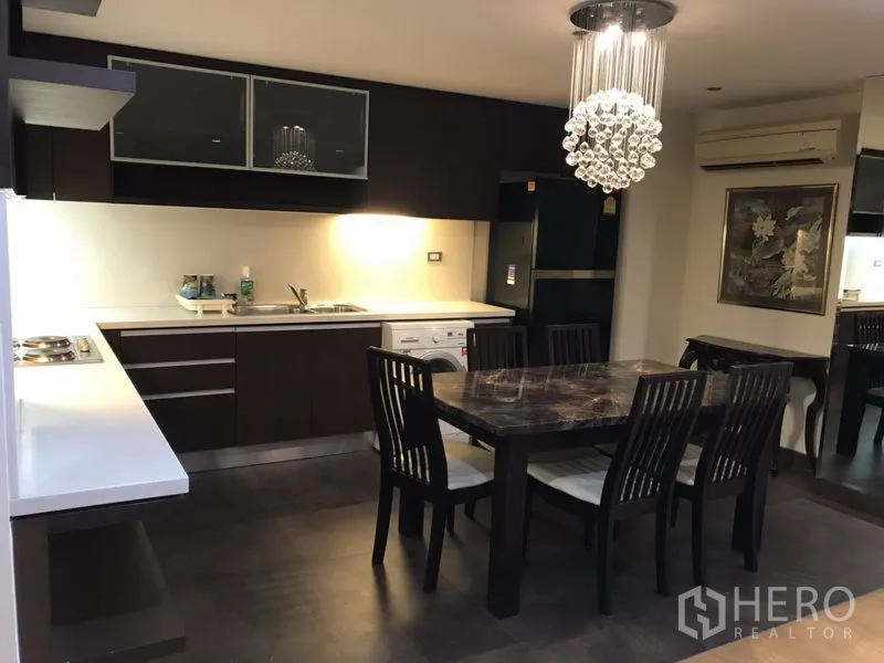 公寓 出租在Watthana, Bangkok - 现代厨房，深色橱柜与白色台面，灶台、洗衣机，餐桌下方水晶吊灯。