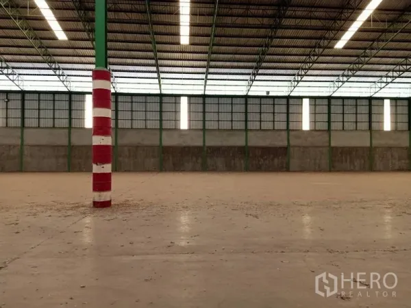 Factory for rent in Chaloem Phra Kiat, Saraburi - Spacious factory interior with high steel truss roof, skylights, concrete floor, and red‑white safety‑striped columns.