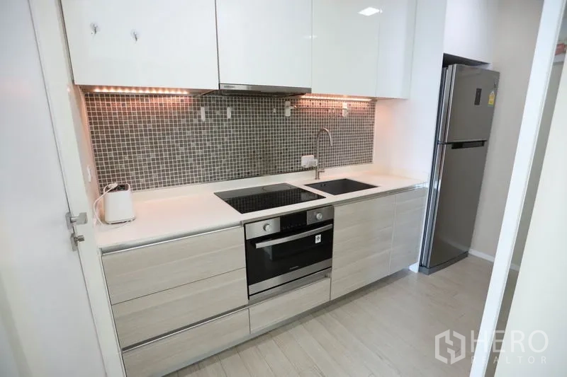 Condominium for rent or sale in Watthana, Bangkok - Contemporary linear kitchen with mosaic backsplash, electric cooktop, oven, sink, and stainless fridge.