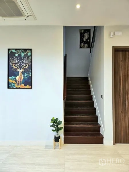 Detached House for rent in Bang Kapi, Bangkok - Dark wood staircase with artwork and potted plant in a white hallway