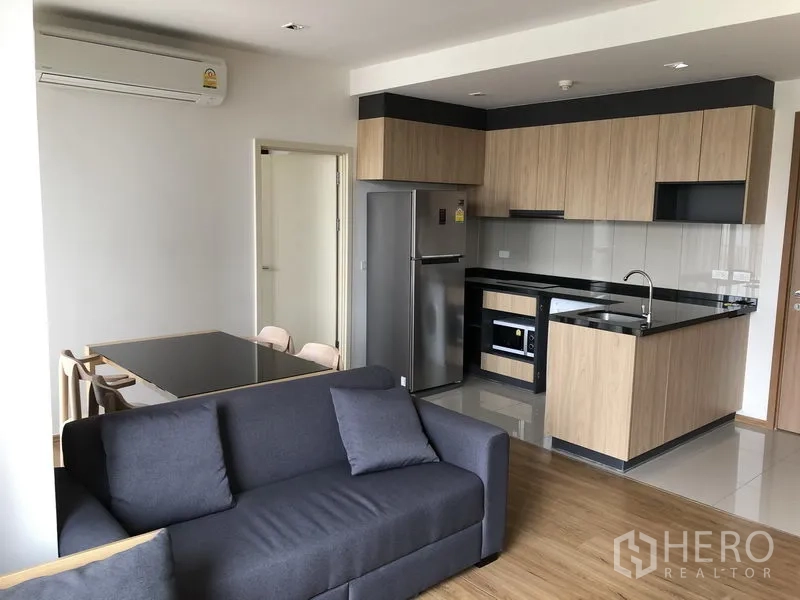 คอนโด ให้เช่าในWatthana, Bangkok - พื้นที่นั่งเล่นแบบเปิดโล่ง โซฟาสีเทา ชุดรับประทานอาหาร และครัวบิ้วท์อินลายไม้ท็อปสีดำพร้อมเครื่องใช้ไฟฟ้า