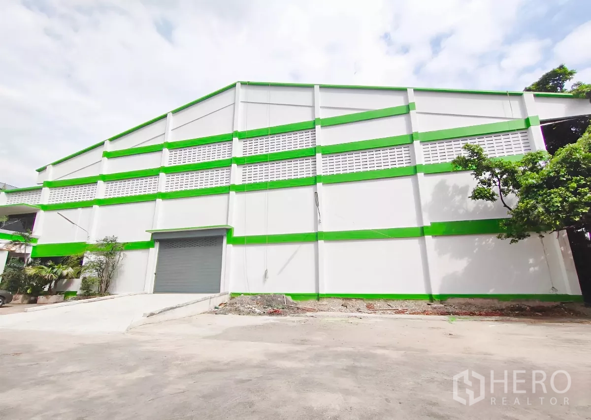 工厂 出售在Samut Prakan - 工厂侧立面，车道与卷闸门，场地周边绿树成荫。