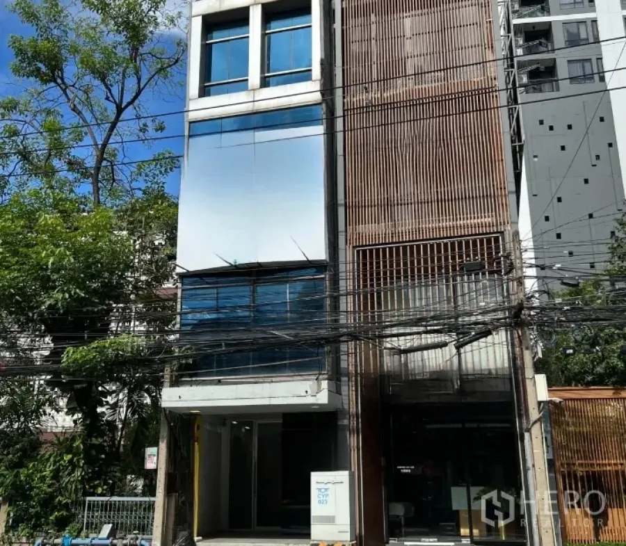 零售空间 出租在Watthana, Bangkok - 四层街铺外立面，玻璃与木质百叶结合，前方有树木与电线。