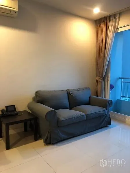 Condominium for sale in Ratchathewi, Bangkok - Living room corner with a gray loveseat, side table and floor-to-ceiling curtains.