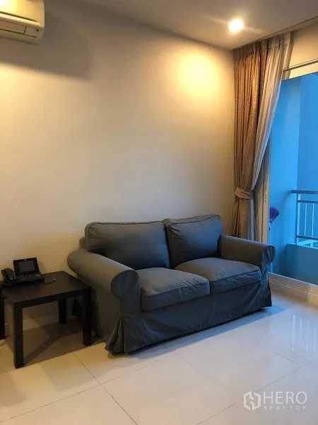 Condominium for sale in Ratchathewi, Bangkok - Living room corner with a gray loveseat, side table and floor-to-ceiling curtains.
