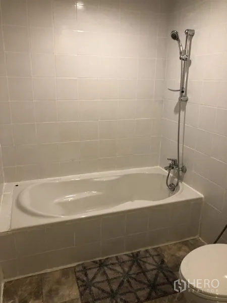 Condominium for rent or sale in Sathon, Bangkok - Bathroom with bathtub and handheld shower against white tiled walls.