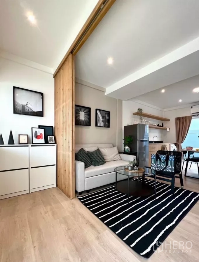 联排别墅 出租在Prawet, Bangkok - 开放式客厅，沙发、条纹地毯、小厨房与木质格栅隔断。