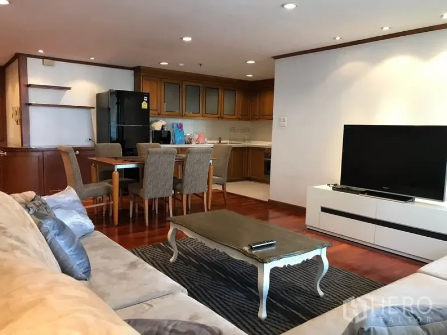 Condominium for rent in Watthana, Bangkok - Open-plan living room with beige sofa, TV console, dining set and wood kitchen cabinetry on hardwood floors.