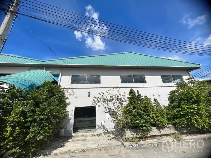 Warehouse for rent in Suan Luang, Bangkok - Warehouse exterior with green metal roof, large windows and landscaped shrubs.