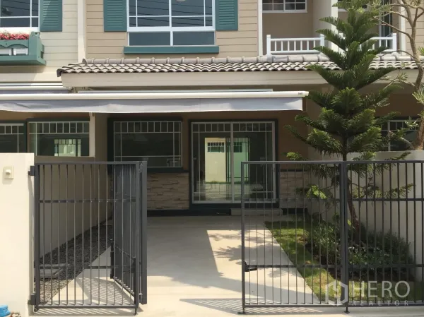 Townhouse for rent in Bang Bo, Samut Prakan - Front view of a two-storey townhouse with gated carport, sliding glass doors and a small lawn.