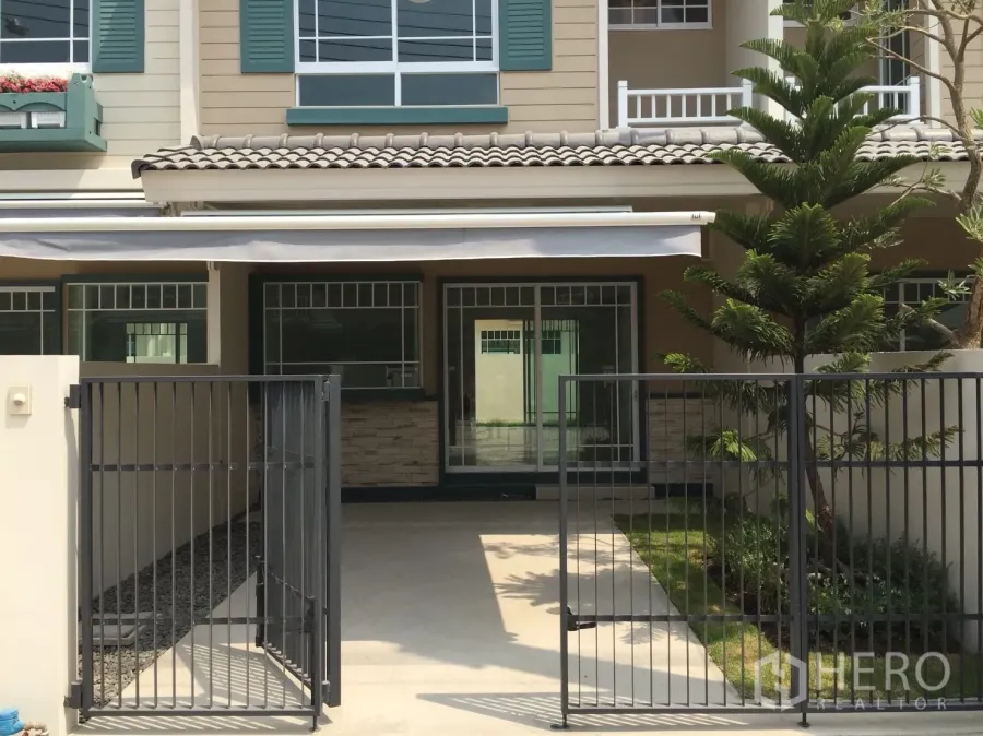 Townhouse for rent in Bang Bo, Samut Prakan - Front view of a two-storey townhouse with gated carport, sliding glass doors and a small lawn.