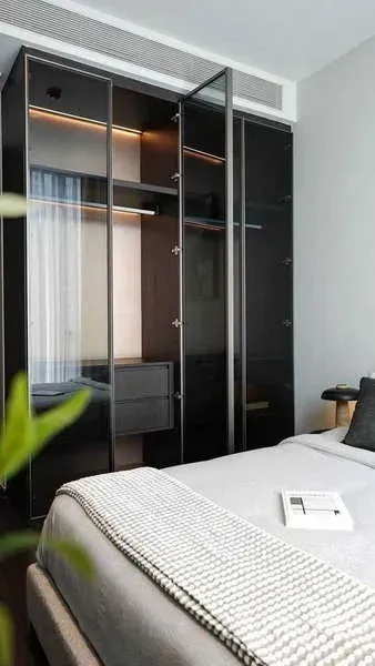 Condominium for rent or sale in Watthana, Bangkok - Bedroom with glass-front wardrobes and soft lighting beside a neatly made bed.