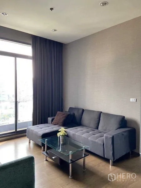 Condominium for sale in Bang Rak, Bangkok - Bright living room with dark sectional sofa, glass coffee table and floor‑to‑ceiling window with city view.