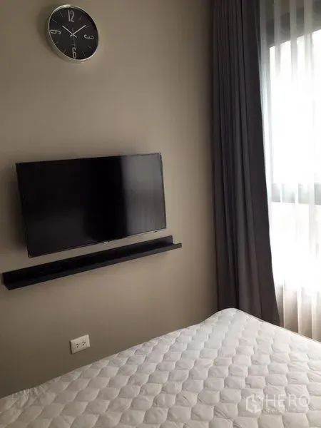 Condominium for sale in Huai Khwang, Bangkok - Wall-mounted flat-screen TV and clock beside a curtained window in the bedroom.