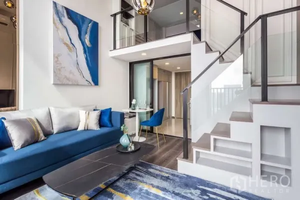 คอนโด ให้เช่า - มุมมองอีกด้านของห้องนั่งเล่นแบบลอฟท์ เห็นบันได ชั้นลอย และโซฟาสีน้ำเงิน