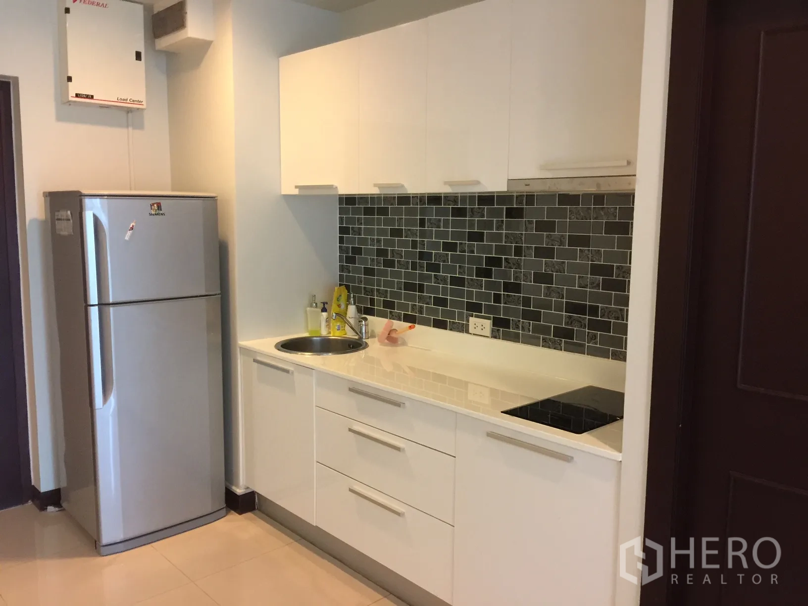 Condominium for rent in Watthana, Bangkok - Compact kitchenette with glossy white cabinets, dark tile backsplash, electric hob and full-size refrigerator.