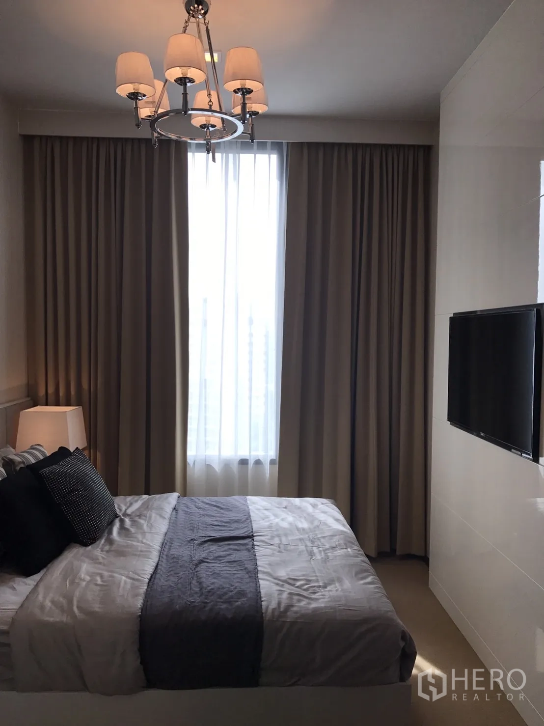 คอนโด ให้เช่าหรือขายในWatthana, Bangkok - มุมห้องนอนมองไปยังหน้าต่างเต็มบาน ผ้าม่านสองชั้น และทีวีติดผนัง ใต้โคมระย้า