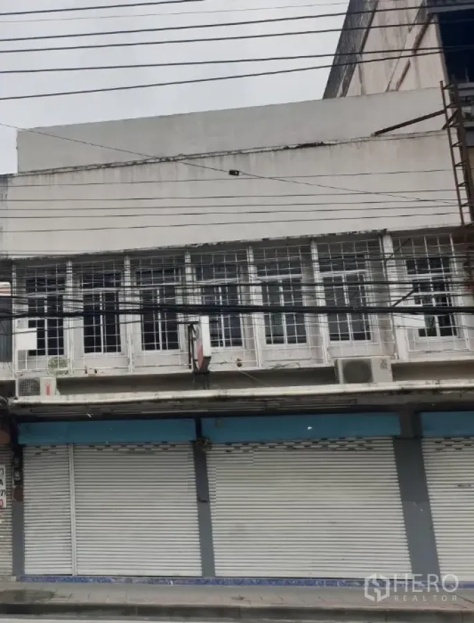 อาคารพาณิชย์ ให้เช่าในWatthana, Bangkok - ด้านหน้าอาคารพาณิชย์ติดถนนในกรุงเทพฯ ประตูม้วนโลหะปิด หน้าต่างชั้นบนแบบกรงเหล็กและสายไฟเหนือถนน.