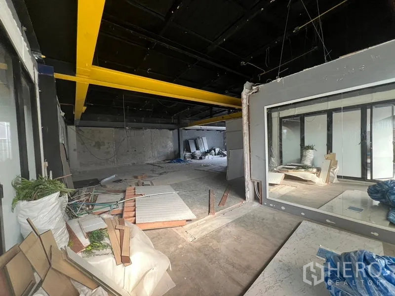 零售空间 出租在Watthana, Bangkok - 室内区域见黄色梁与大内窗分隔，地面有施工碎料。