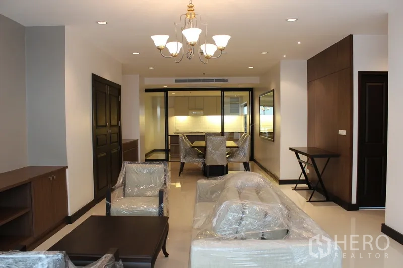 Condominium for rent in Watthana, Bangkok - Open-plan living and dining area with chandelier lighting, protected new furniture and view to the glass-enclosed kitchen.