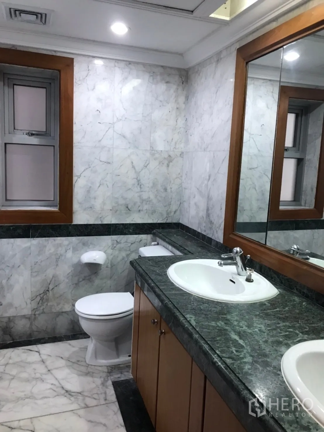 Condominium for rent or sale in Pathum Wan, Bangkok - Bathroom with marble walls, long stone countertop, sink, large mirror and toilet.