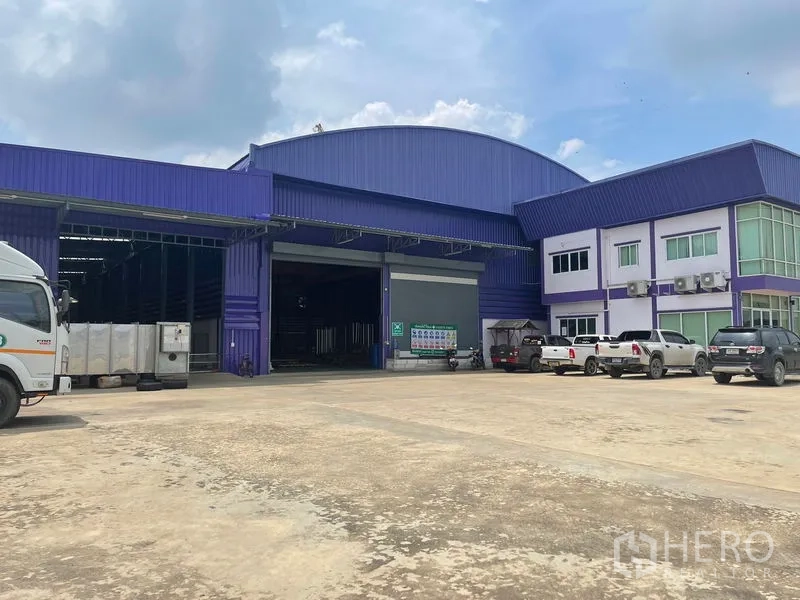Warehouse for sale in Lam Luk Ka, Pathum Thani - Front view of a large purple-and-blue warehouse with high arched roof, wide roller shutter doors, attached two-storey glass-corner office and a spacious concrete yard with parked trucks.