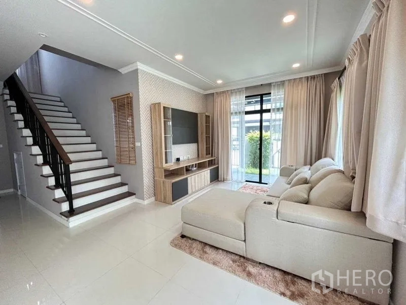 Detached House for rent in Khlong Sam Wa, Bangkok - Light-filled living room with L-shaped sofa, media wall, staircase and sliding doors to the garden
