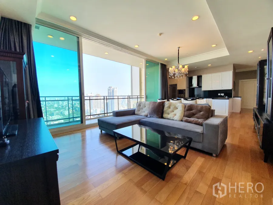 公寓 出租在Watthana, Bangkok - 采光充足的客厅，L形沙发与玻璃茶几，落地窗通往高层阳台，俯瞰城市天际线。