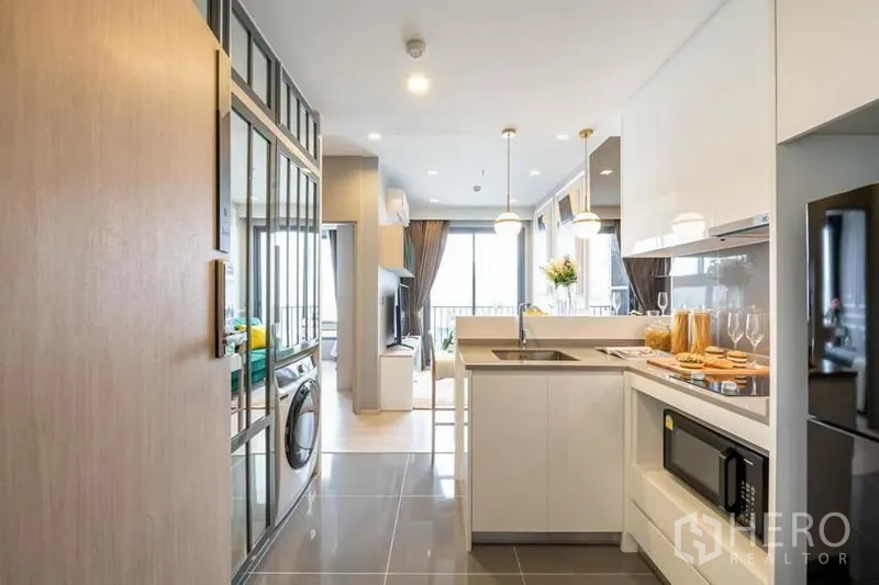 Condominium for rent or sale in Watthana, Bangkok - Sleek L-shaped kitchen with built-in appliances, pendant lighting and outlook to the living area, plus washing machine.