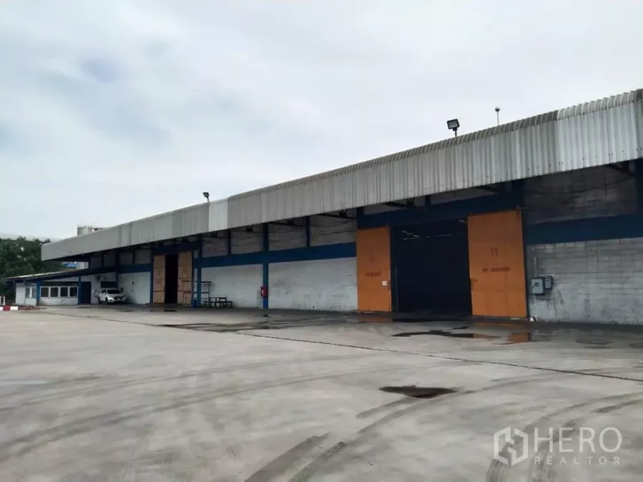 工厂 出租在Samut Prakan - 工厂外立面设有宽阔混凝土场地、带顶装卸区与大型推拉门，卡车进出便捷，位于帕巴登。
