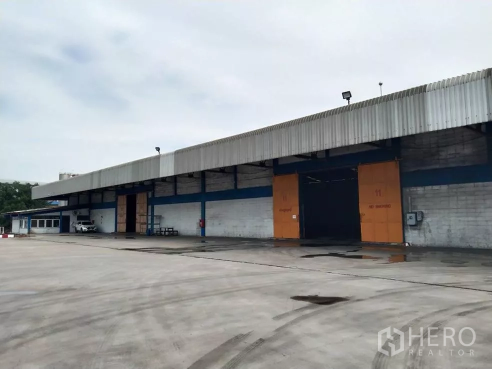 工厂 出租在Samut Prakan - 工厂外立面设有宽阔混凝土场地、带顶装卸区与大型推拉门，卡车进出便捷，位于帕巴登。