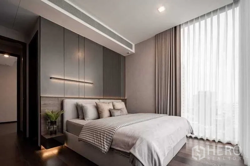คอนโด ให้เช่า - ห้องนอนหลัก หน้าต่างสูงจรดเพดาน ผนังพาเนลสีเทา ไฟซอฟท์ และวิวเมืองผ่านม่านบาง