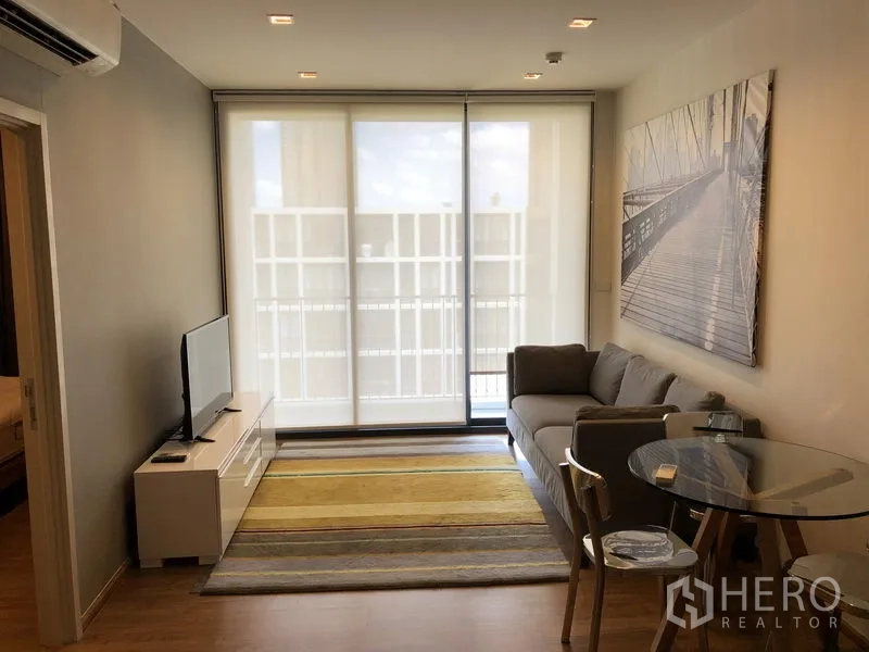 Condominium for rent in Watthana, Bangkok - Bright living room with gray sofa, area rug, TV console and floor-to-ceiling windows opening to a balcony.