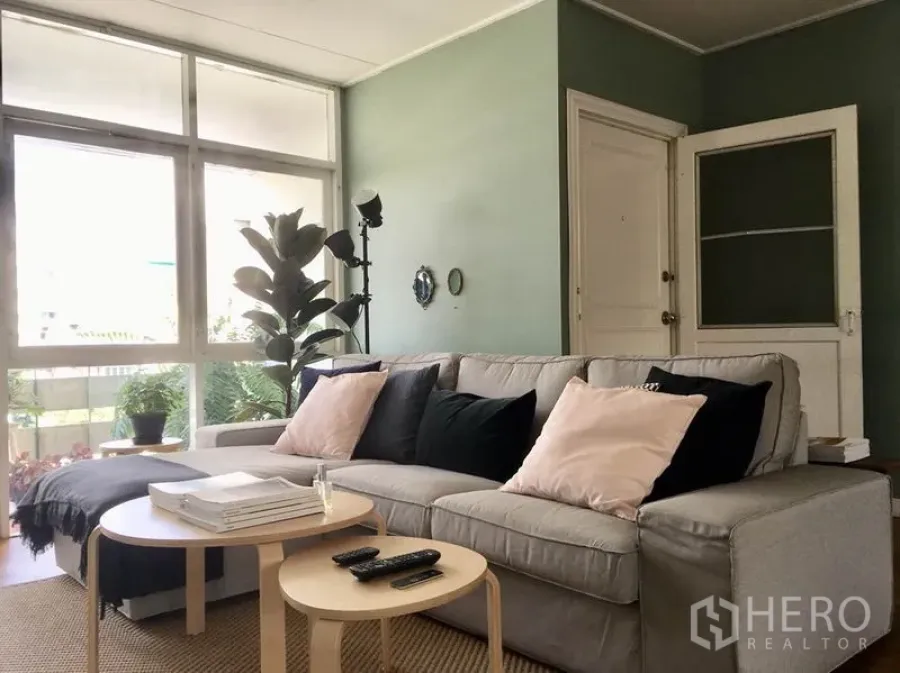 Apartment for rent in Khlong Toei, Bangkok - Bright living room with gray sofa, nesting tables, and floor-to-ceiling windows overlooking greenery in a Thonglor Bangkok apartment.