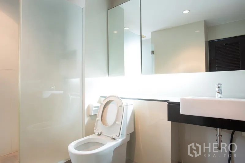 Condominium for rent in Watthana, Bangkok - Modern bathroom with glass shower, toilet, and long illuminated mirror.