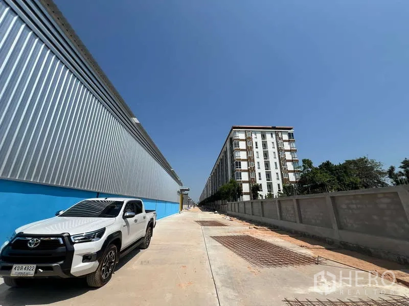 货栈 出租在Lat Krabang, Bangkok - 项目道路与现场停车位，临金属外墙仓库，旁有低层住宅楼。